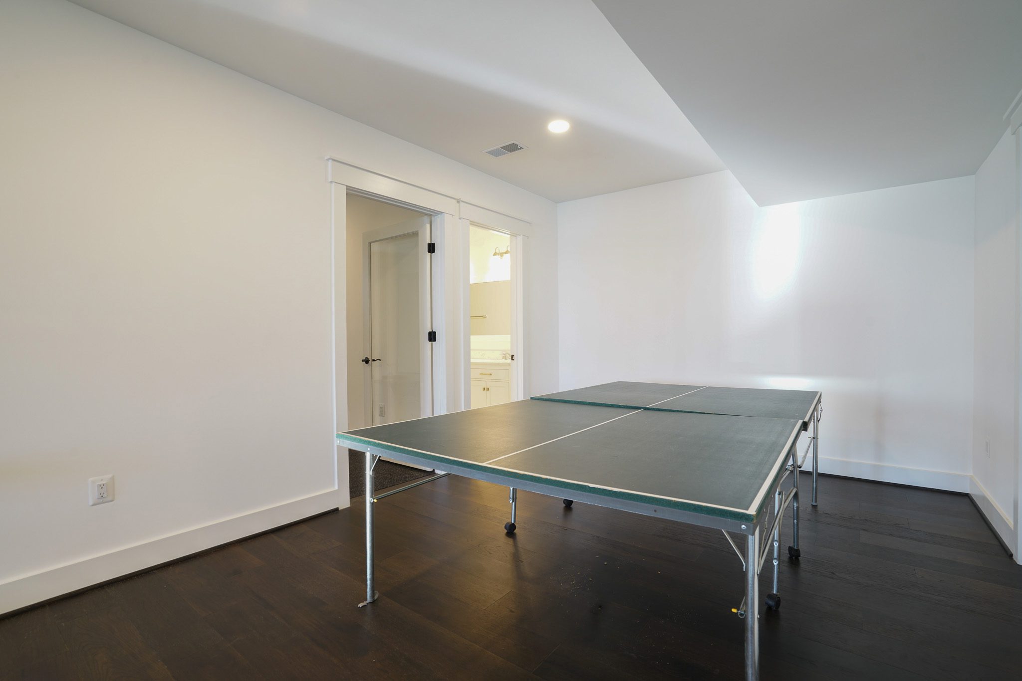 Room with a ping pong table on a dark wood floor, white walls, and a door leading to a bathroom.