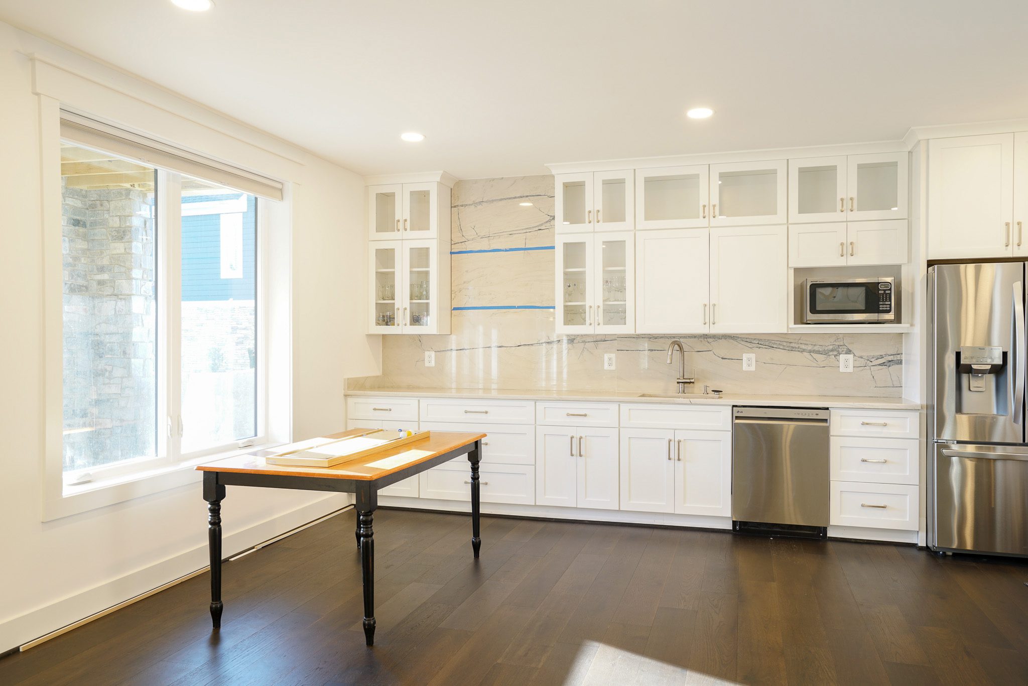 Bright kitchen with white cabinets, stainless steel appliances, a wooden table, and a large window. Marble backsplash with light and shadow contrast.