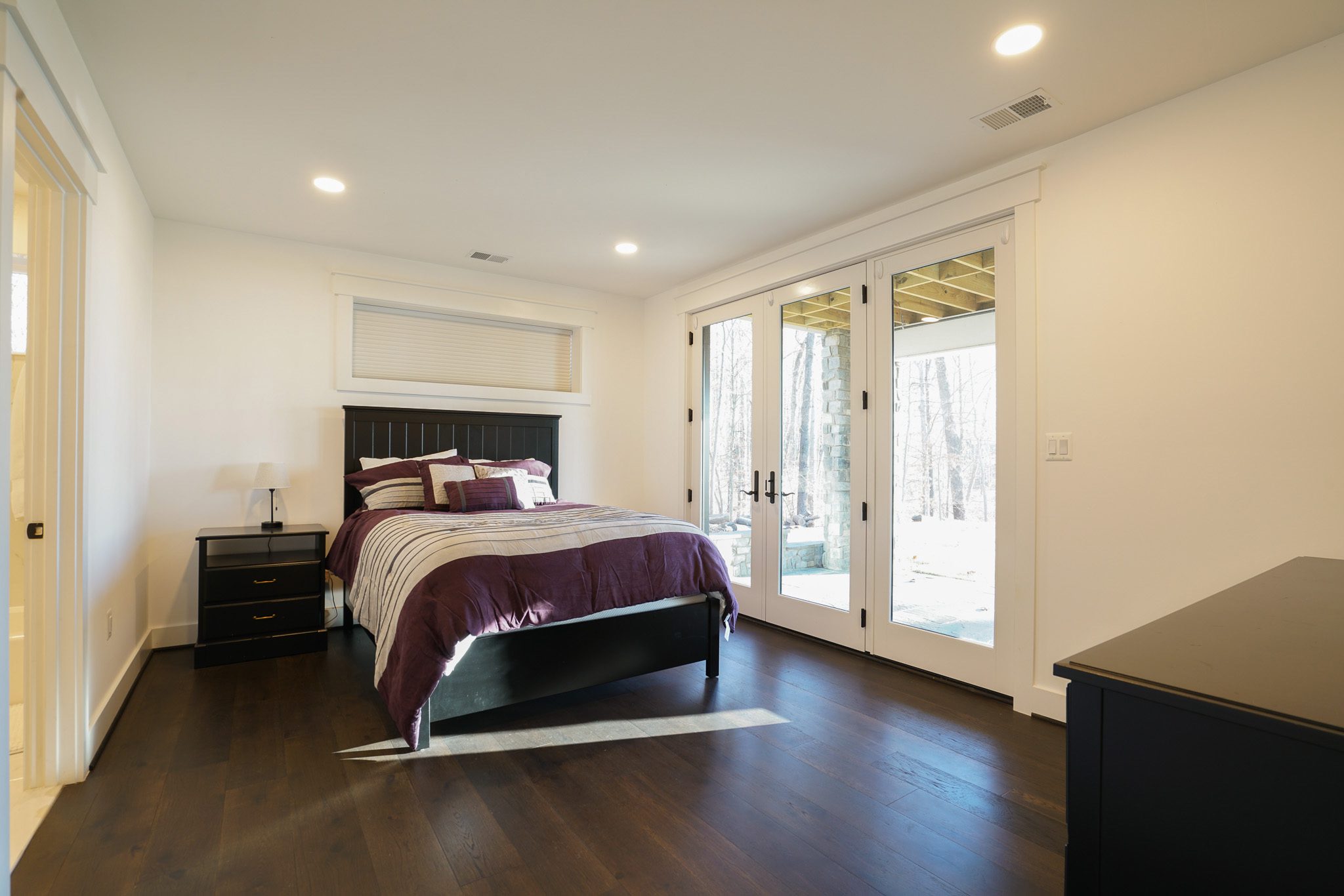 A bedroom with a double bed, dark wood floor, bedside table with lamp, and large glass doors leading to an outdoor area.
