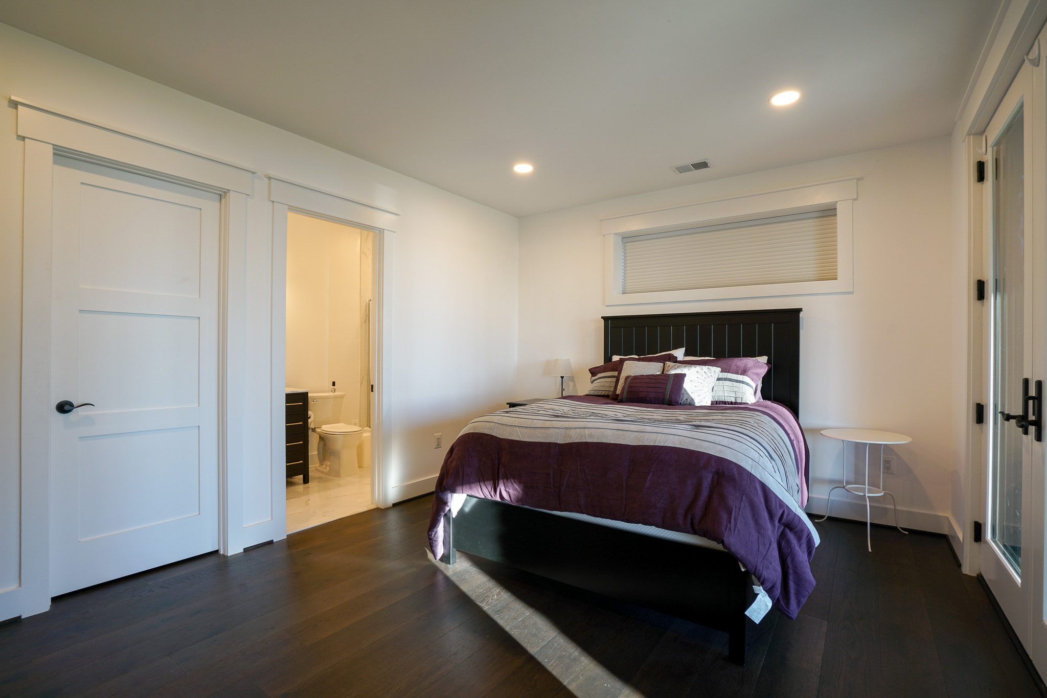 Bedroom with a double bed dressed in purple and gray bedding, a small white side table, wood flooring, and an open door leading to a bathroom.