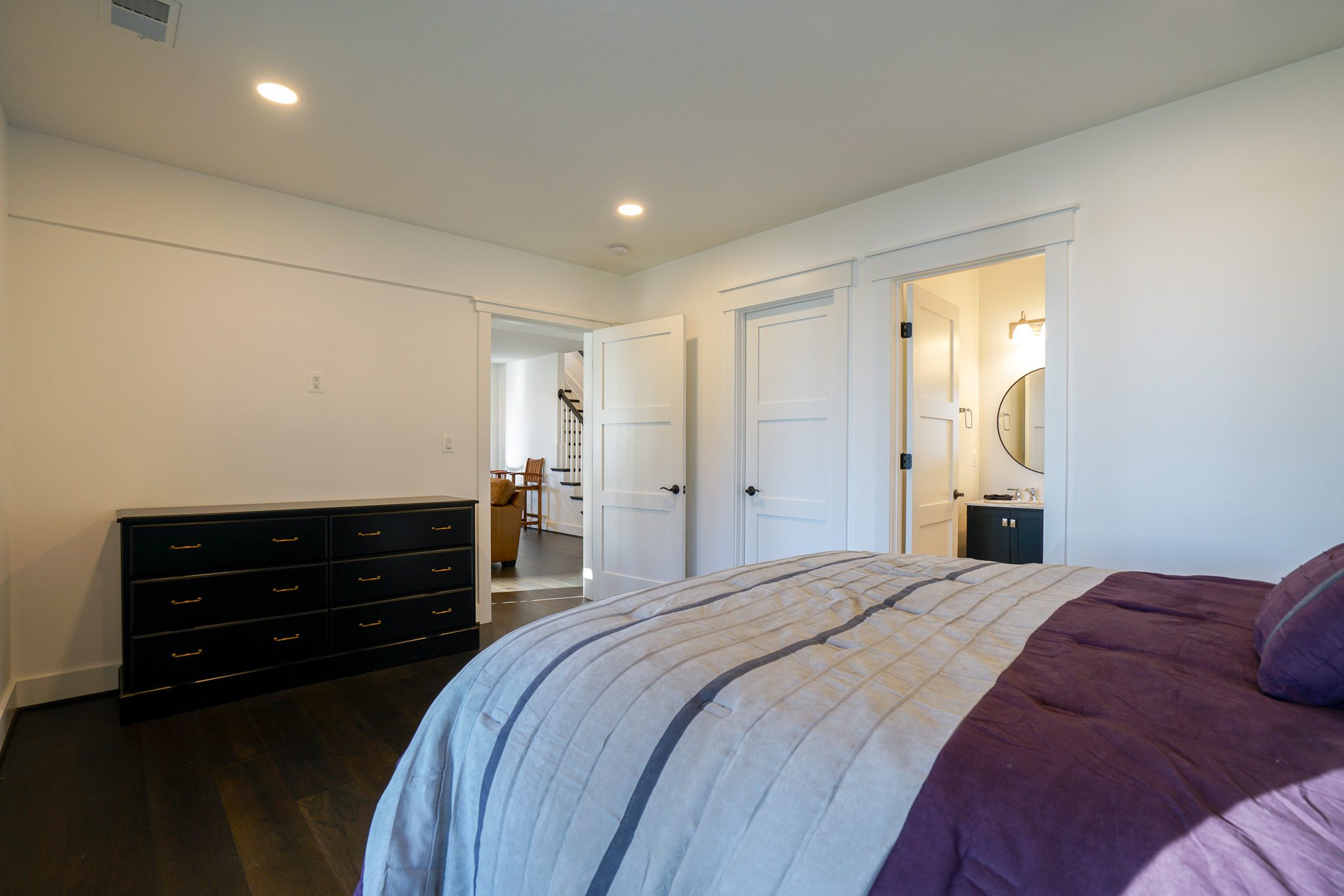 A bedroom with a bed, dark wood dresser, and open doorways leading to a bathroom and hallway. White walls, dark flooring, and a ceiling light are visible.