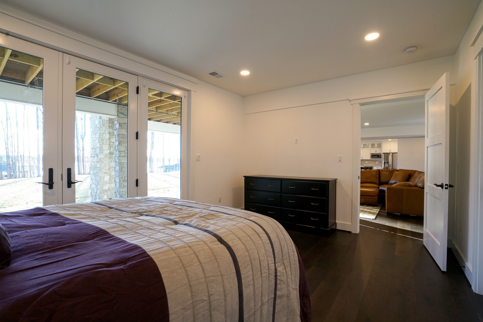 Bedroom with a bed, striped bedding, dark wood floor, and a dresser. French doors open to an outdoor area. An open door shows a glimpse of a living area with a brown couch.