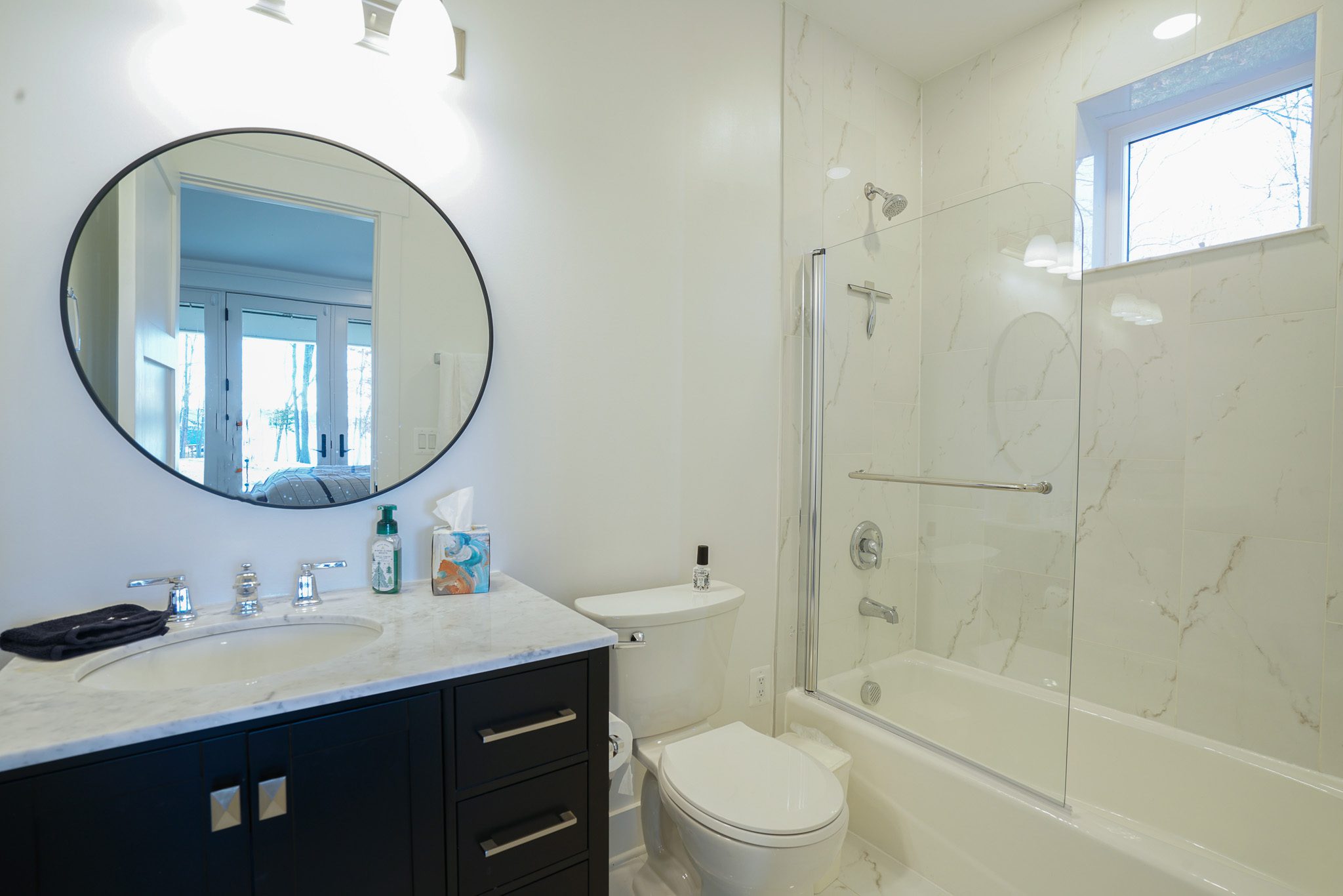 Modern bathroom with a round mirror above a black vanity, marble countertop, and a toilet. There is a shower with glass doors and a small window above. Reflections show a bright room.