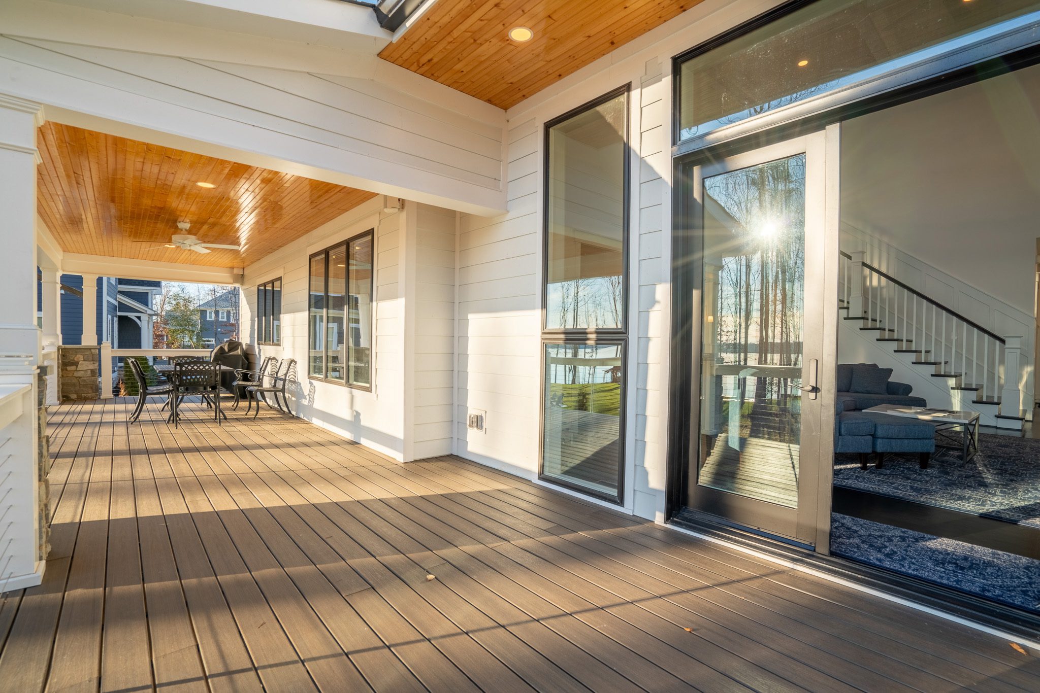 Spacious porch with wooden flooring, outdoor dining area, and large glass doors leading to an interior room with a staircase. Sunlight streams through the glass.