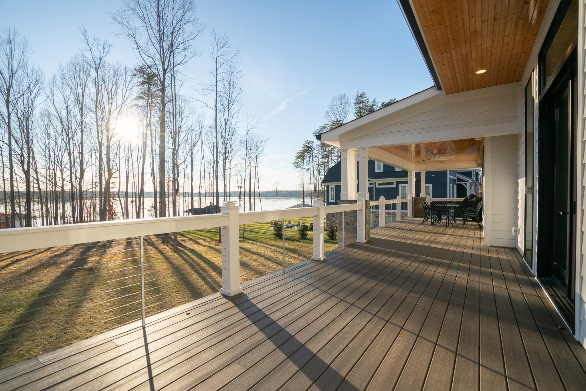 Large wooden deck on a modern house, overlooking a grassy yard with bare trees and a lake in the background under a clear sky with the sun low on the horizon.