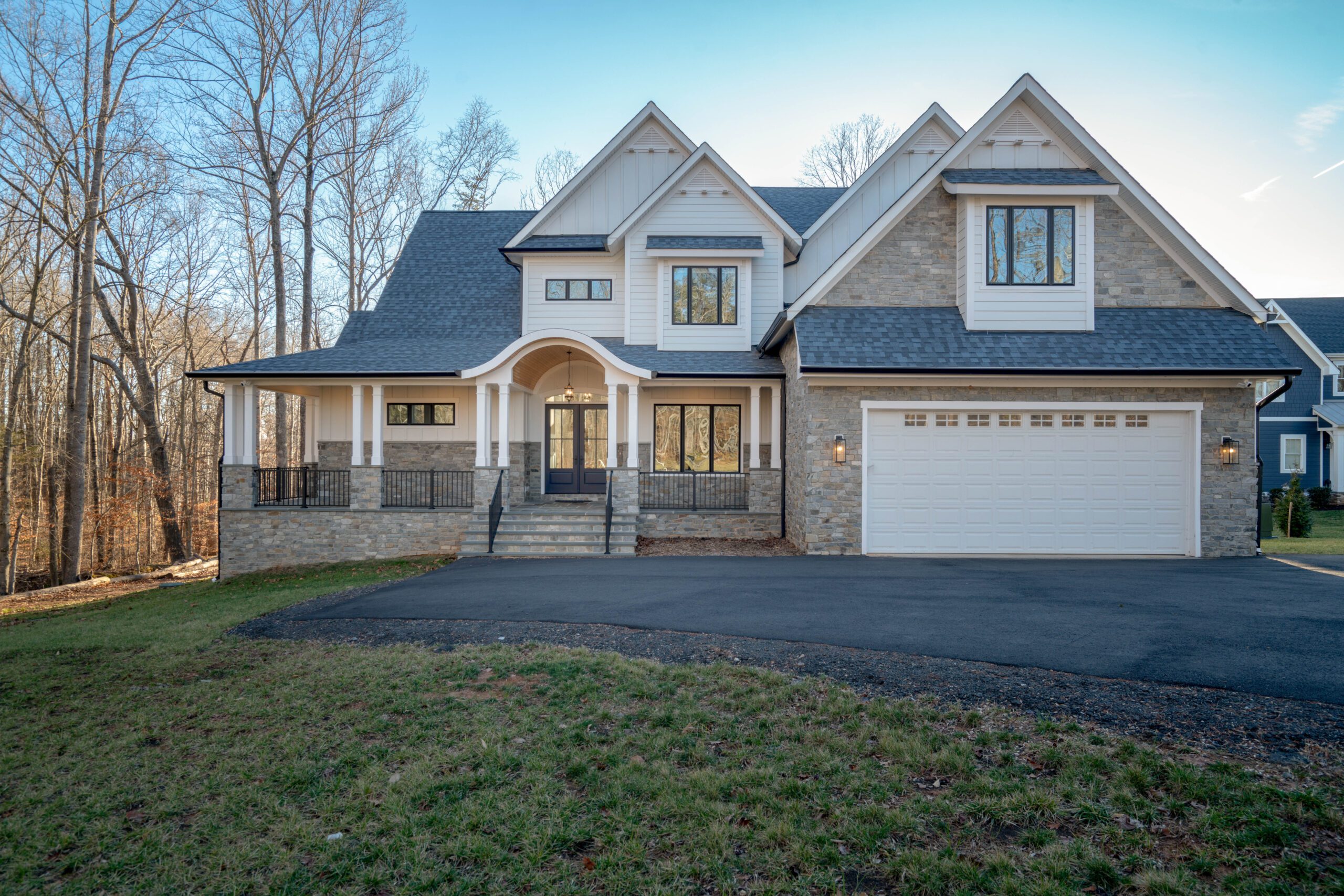 Two-story house with stone and siding exterior, large front porch, and three-car garage, set against a bare tree backdrop.