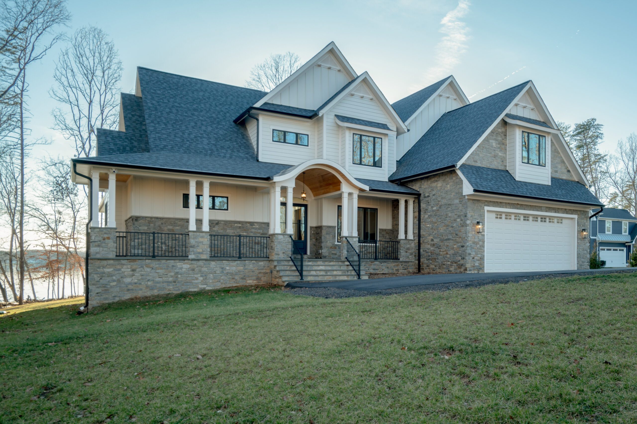 A large modern house with a stone and wood exterior, featuring multiple gables and a spacious front porch, sits on a grassy lawn with trees and a glimpse of water in the background.