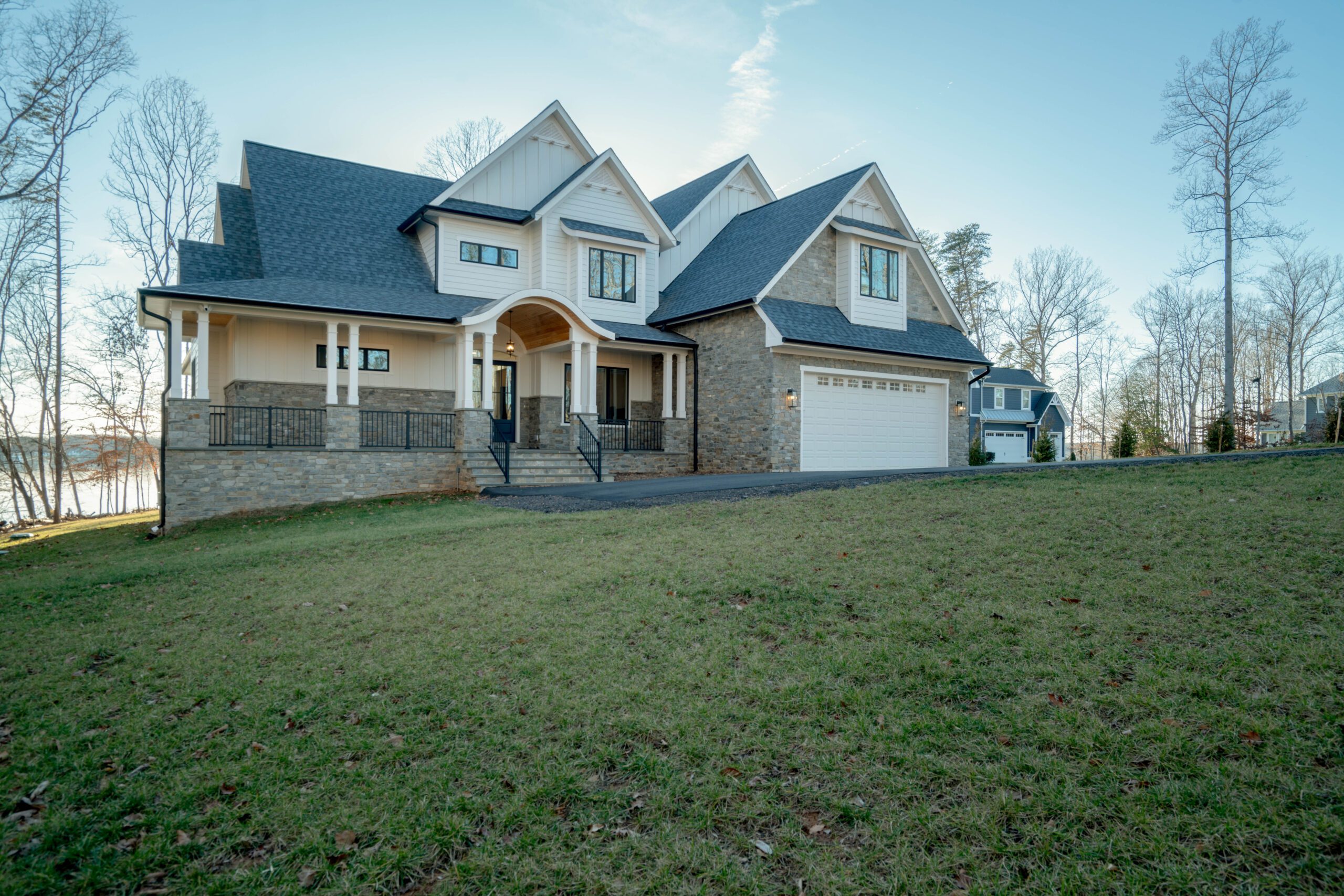 A two-story house with a stone and siding exterior, dark shingled roof, and attached garage, set on a grassy lawn with bare trees and a view of the water in the background.