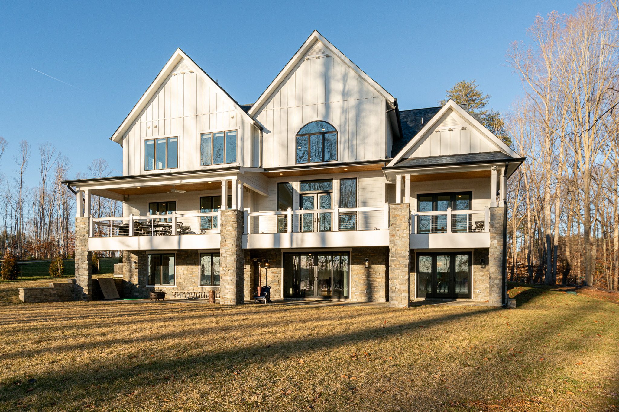 Two-story modern house with large windows, stone columns, and multiple balconies, set on a spacious lawn with surrounding trees under a clear blue sky.