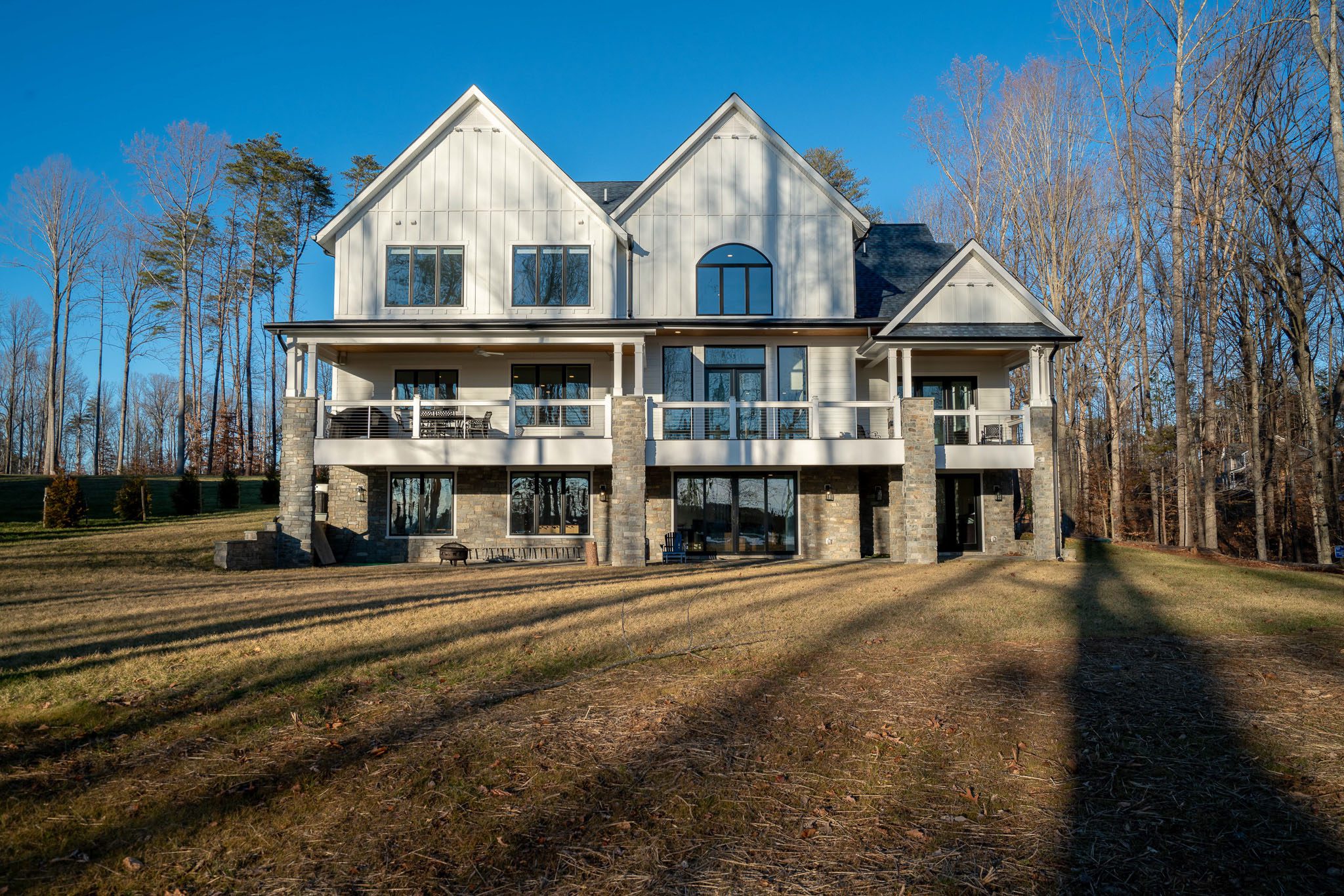 A modern, multi-story house with large windows, set in a wooded area on a sunny day. The lawn in front is expansive, with trees visible in the background.
