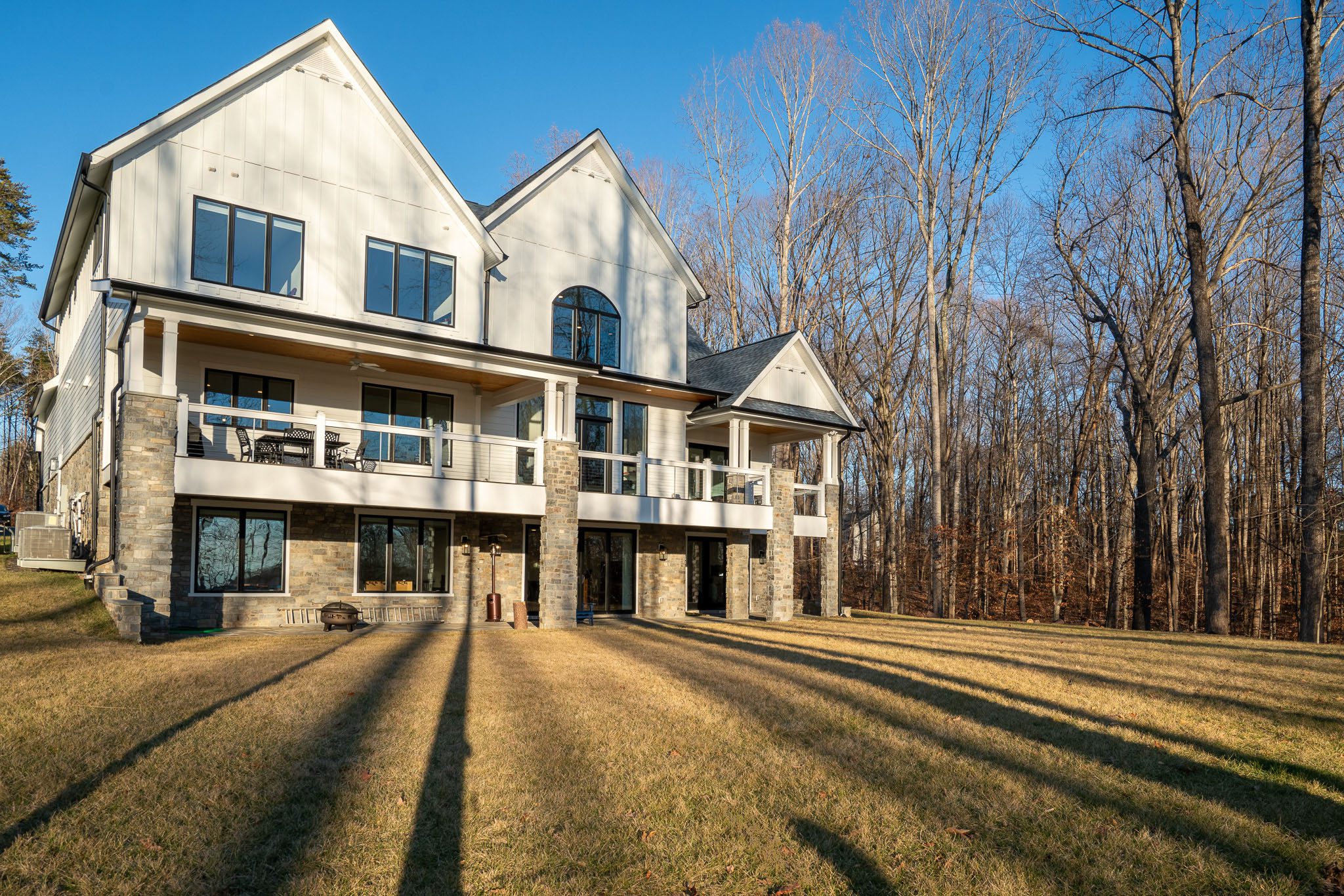 Large modern house with multiple balconies, white walls, and stone accents, surrounded by grass and bare trees under a clear blue sky.