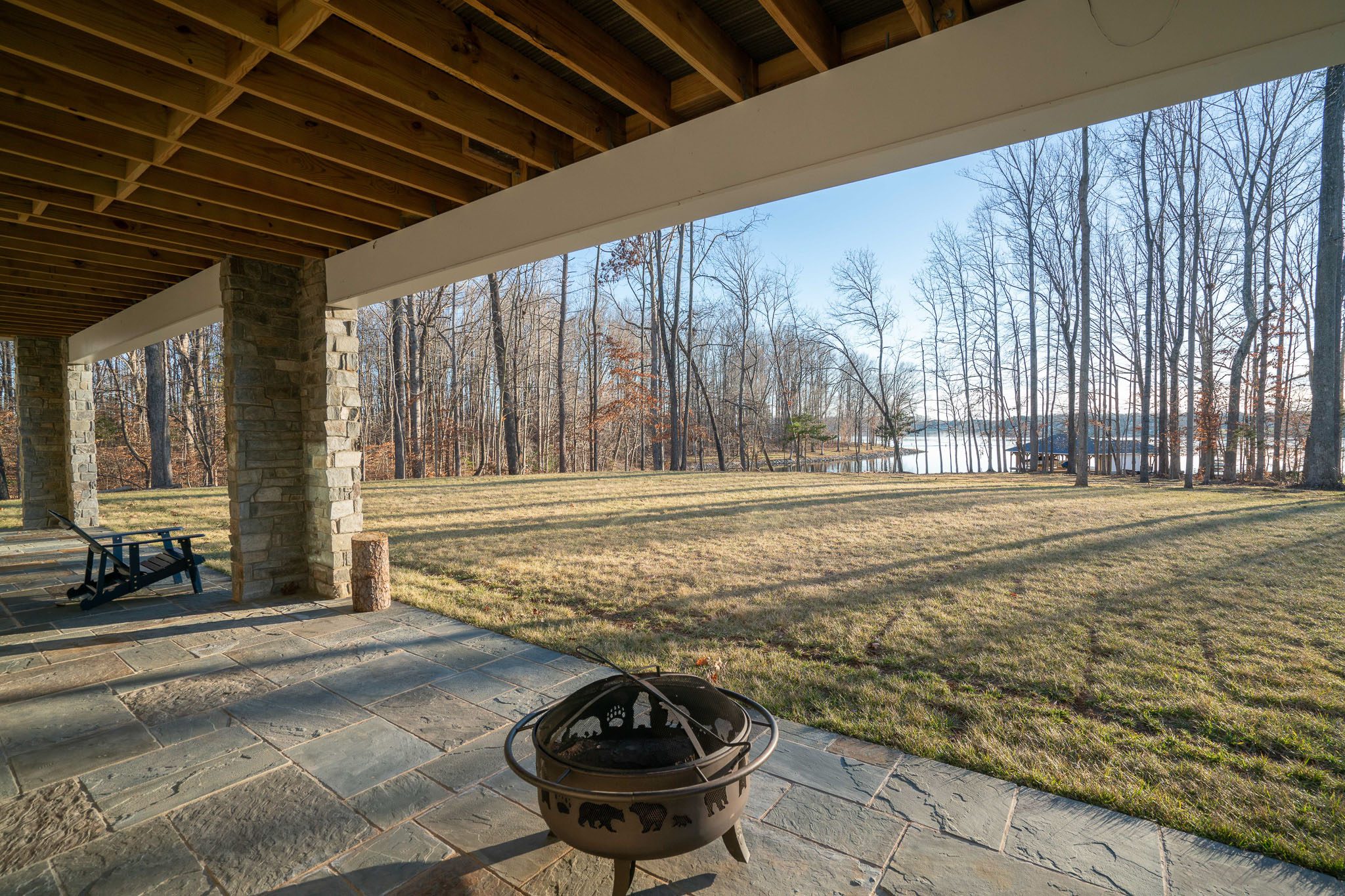 Covered patio with stone flooring, a fire pit, and a view of a grassy yard leading to a lake bordered by leafless trees.