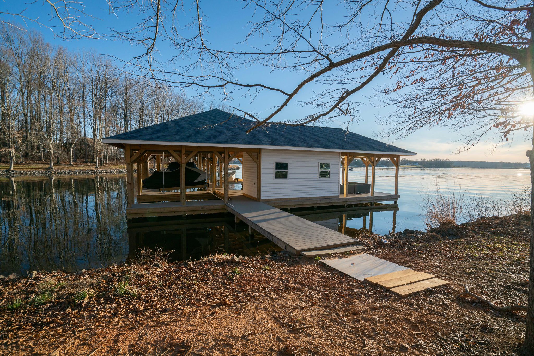 A wooden dock leads to a covered boathouse on a calm lake, surrounded by bare trees under a clear blue sky.