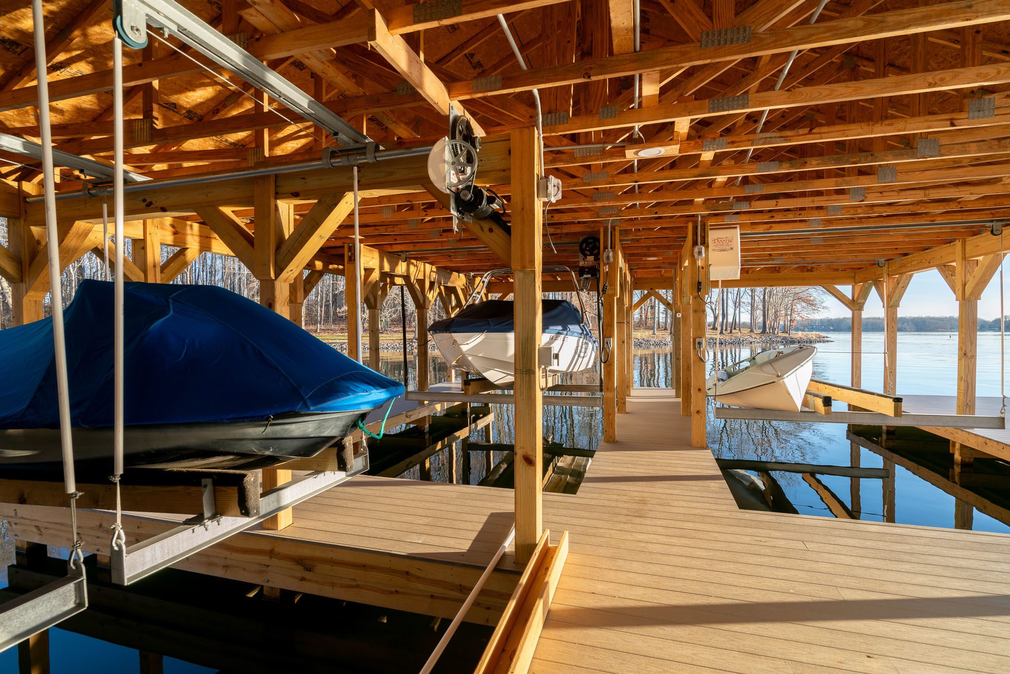 A covered wooden dock with boats secured inside. Two boats are visible in the foreground, one covered in blue. Sunlight illuminates the wooden beams and the water is calm.