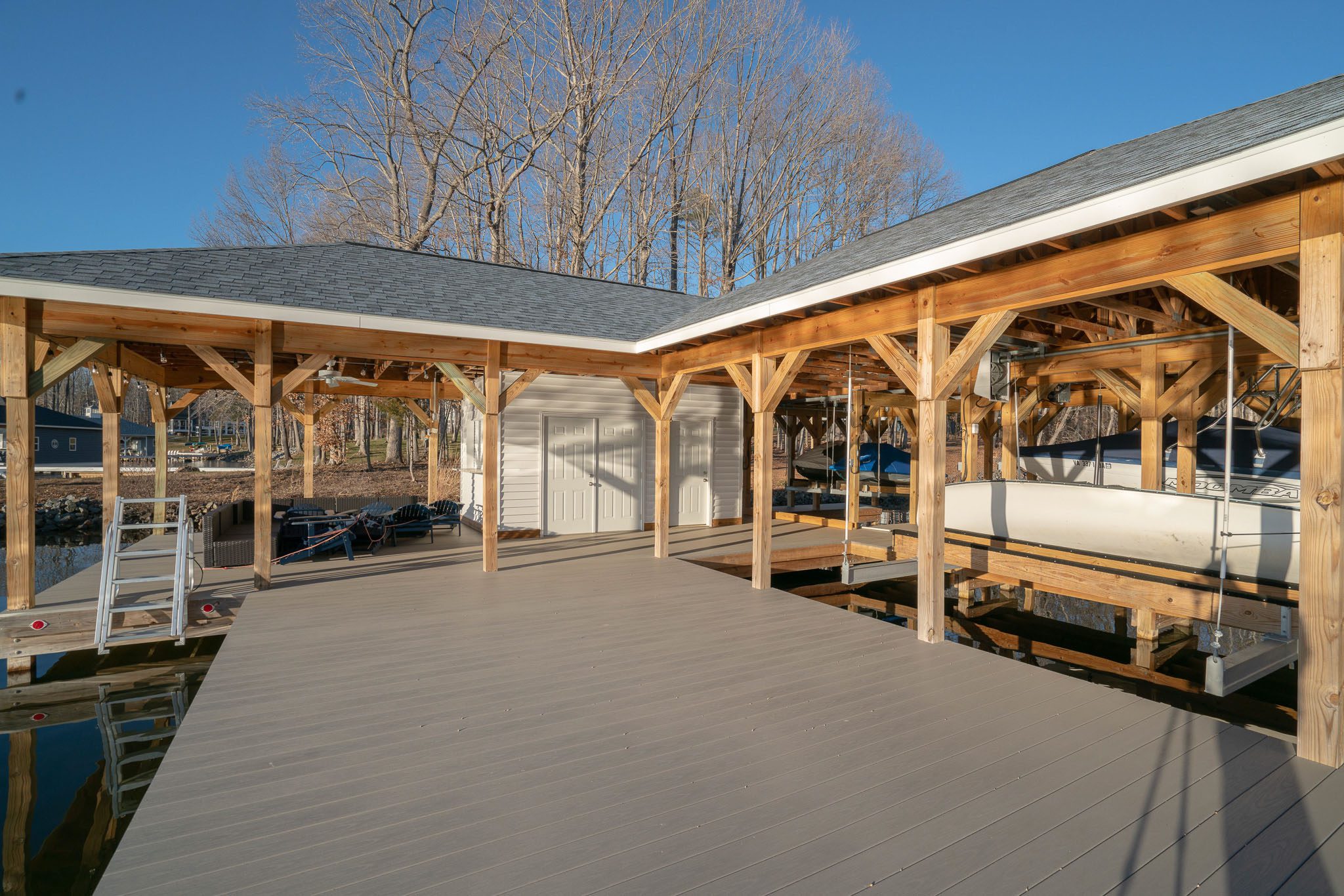 Wooden dock structure with a boat in a sheltered area, surrounded by bare trees. The dock has a sloped roof and is situated on calm water on a clear day.