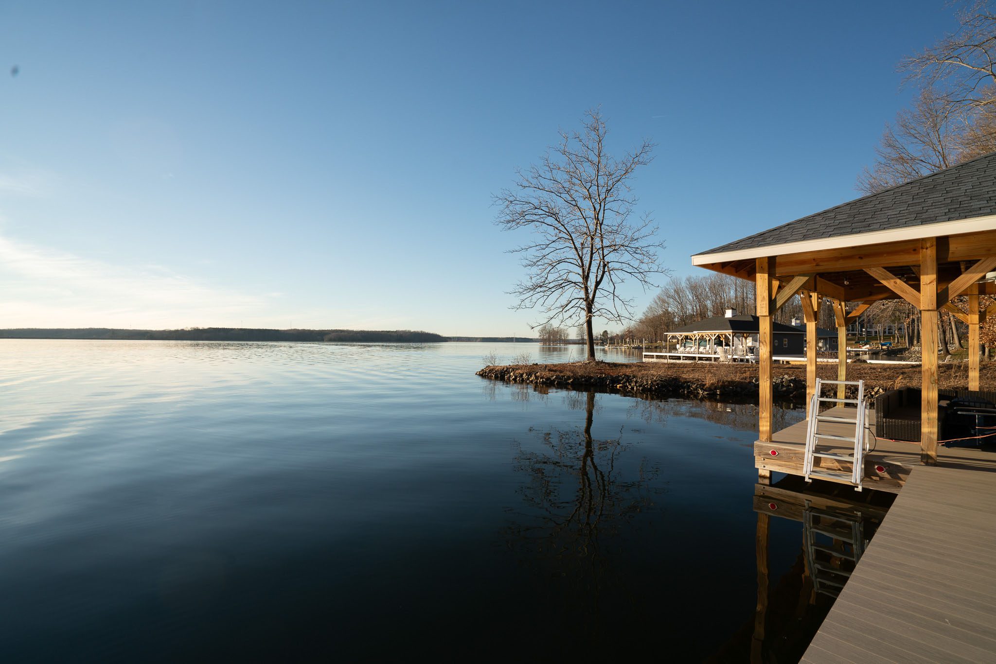 Lakeside view with a wooden dock, a leafless tree, and calm water under a clear blue sky.