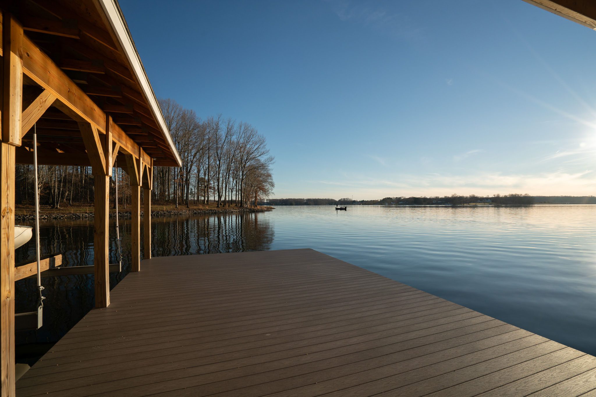 Wooden dock on a calm lake with a small boat visible in the distance, set against a clear blue sky and bare trees along the shoreline.