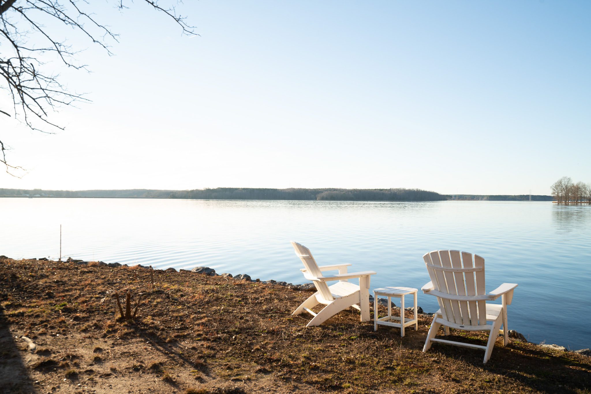 Two white wooden chairs face a calm lake with clear skies. Sparse trees are visible on the opposite shore.
