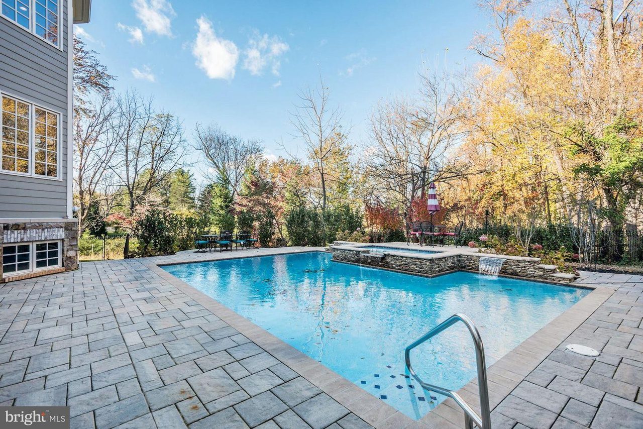 A rectangular outdoor swimming pool surrounded by stone tiles, next to a house. Trees with autumn foliage are in the background under a blue sky.