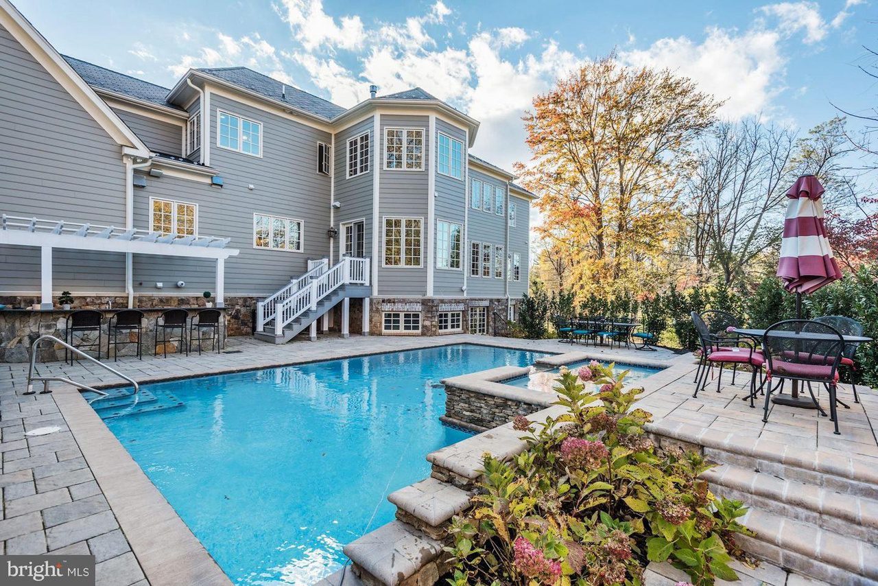 Large house with a backyard swimming pool and adjacent patio. The area features outdoor seating, a pergola, and autumn trees in the background.