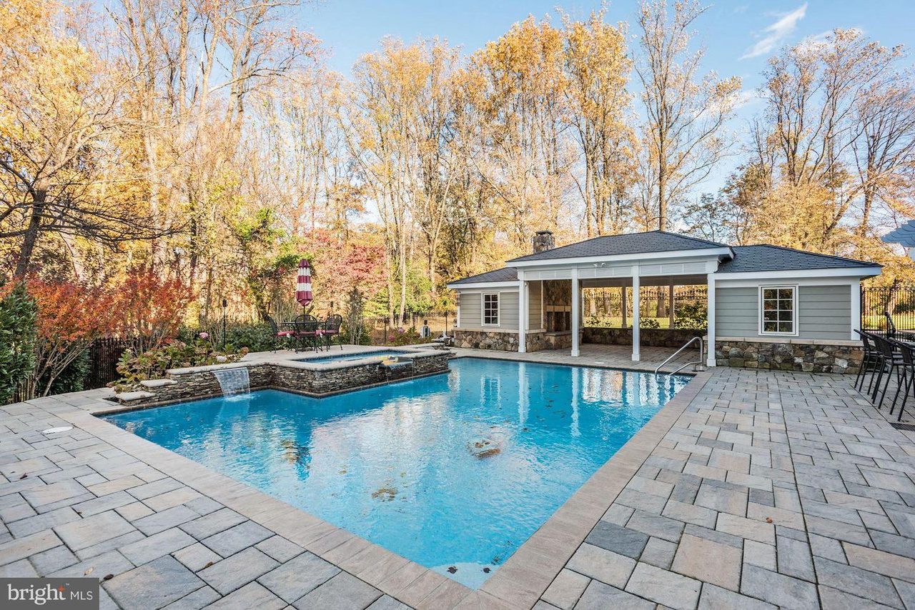 Outdoor pool with a connected spa, surrounded by stone tile decking, next to a small pool house. Autumn trees fill the background.