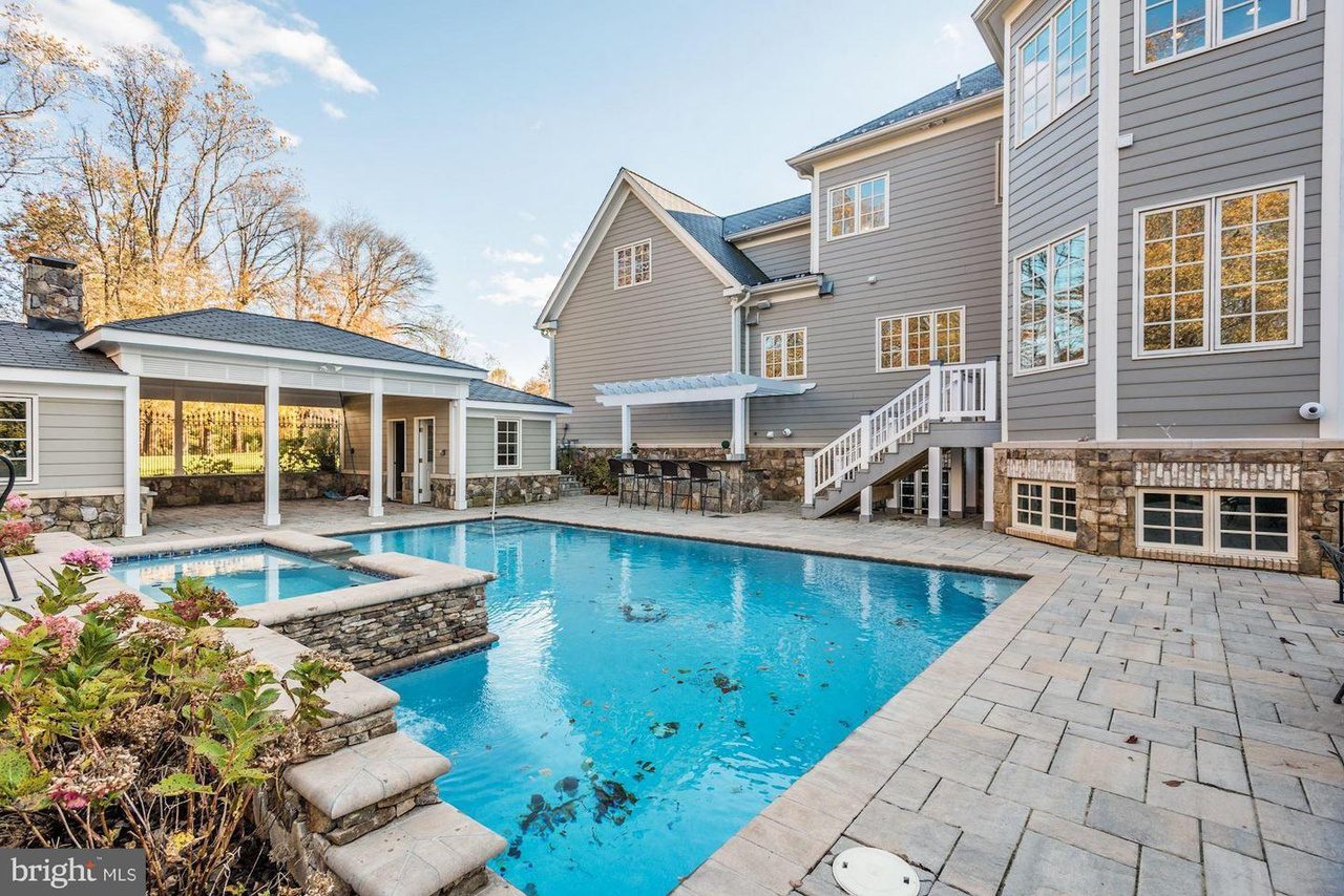 Large house with multiple stories, a backyard pool, and a stone patio. White pergola and fence surround the area. Trees and blue sky are visible in the background.