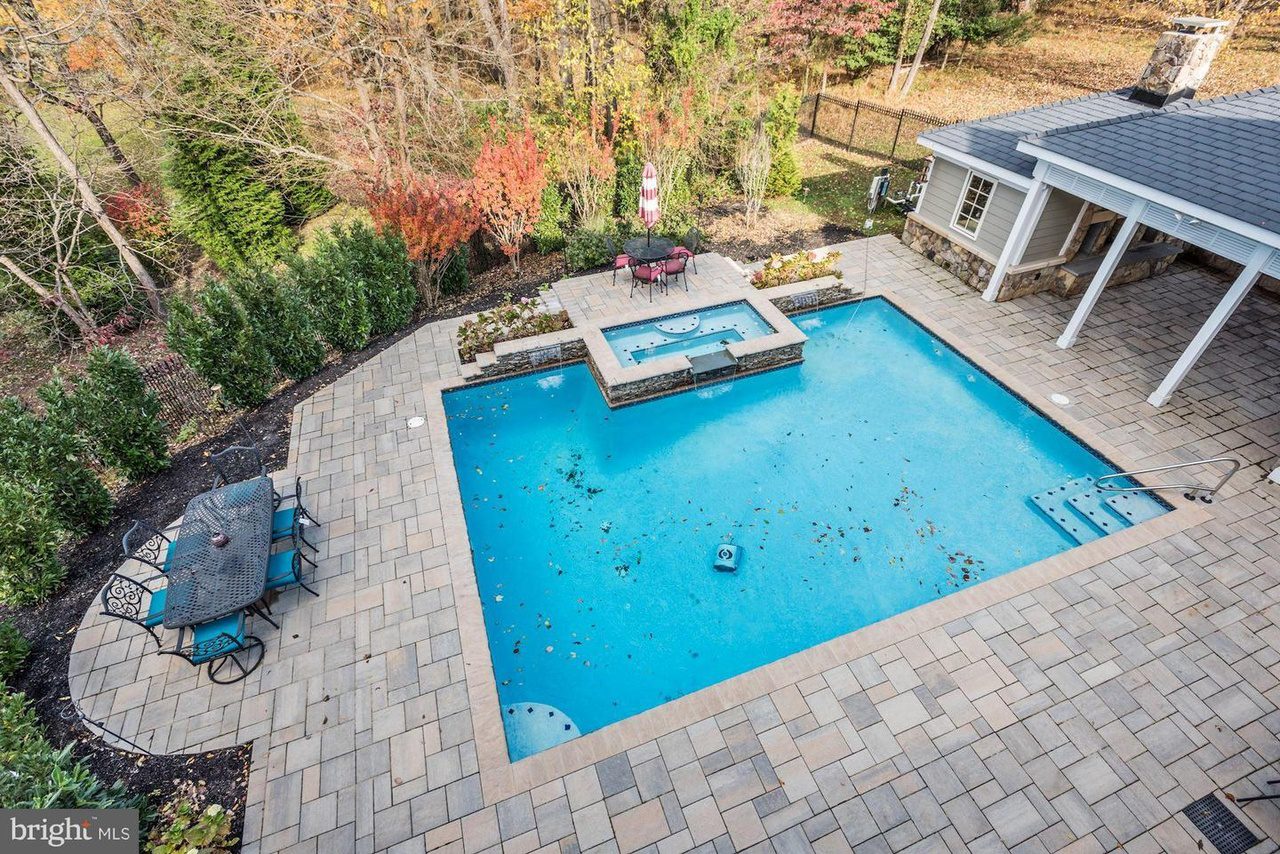 Aerial view of a rectangular outdoor pool with nearby seating areas and trees in the background. The patio is paved with light-colored tiles.
