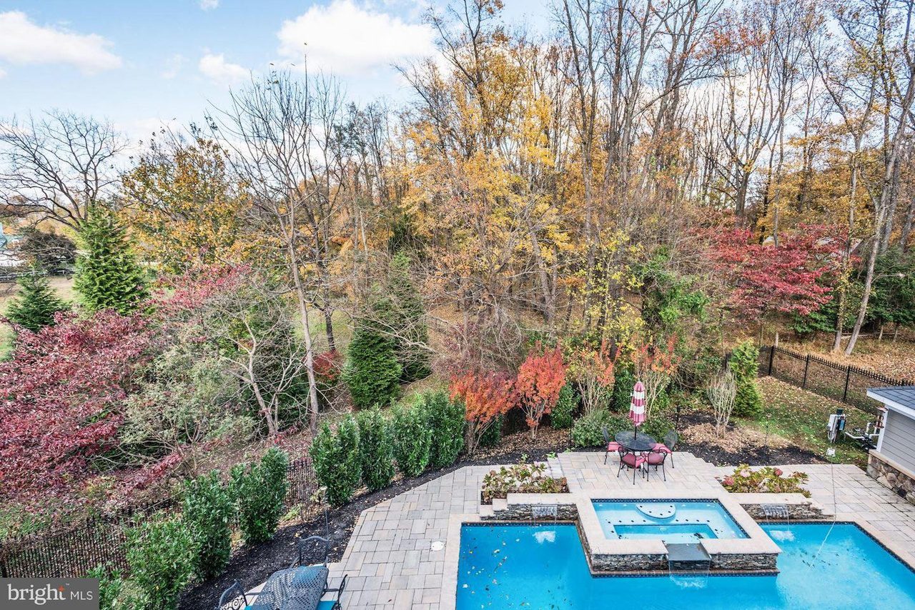 A backyard scene with a swimming pool, patio, and outdoor seating area surrounded by trees with autumn foliage and manicured bushes.