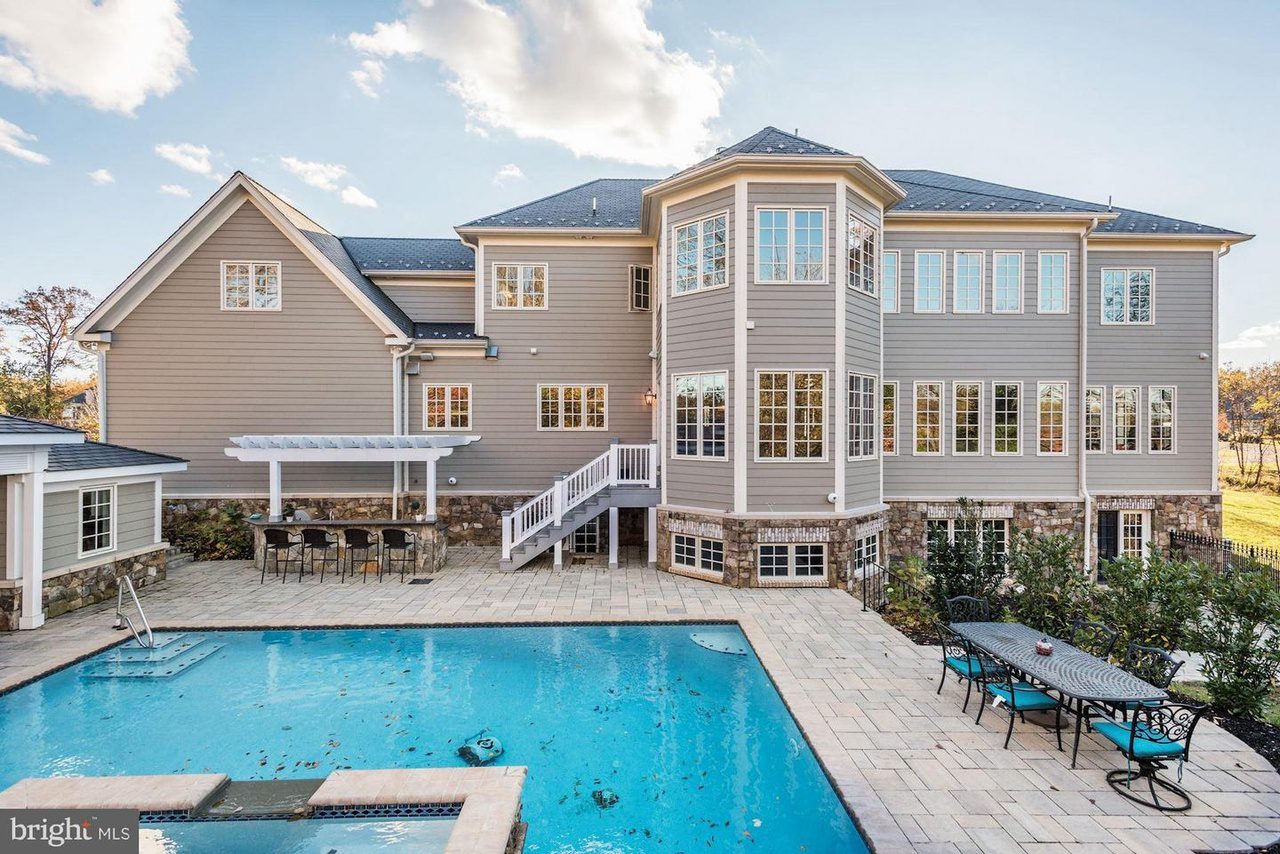 Large multi-story house with gray siding, numerous windows, and a stone foundation. The backyard features a swimming pool, a hot tub, and a patio with outdoor tables and chairs.