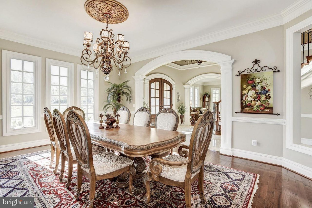Elegant dining room with a long wooden table, ornate chairs, a chandelier, and large windows. Features an archway leading to another room and a colorful floral painting on the wall.