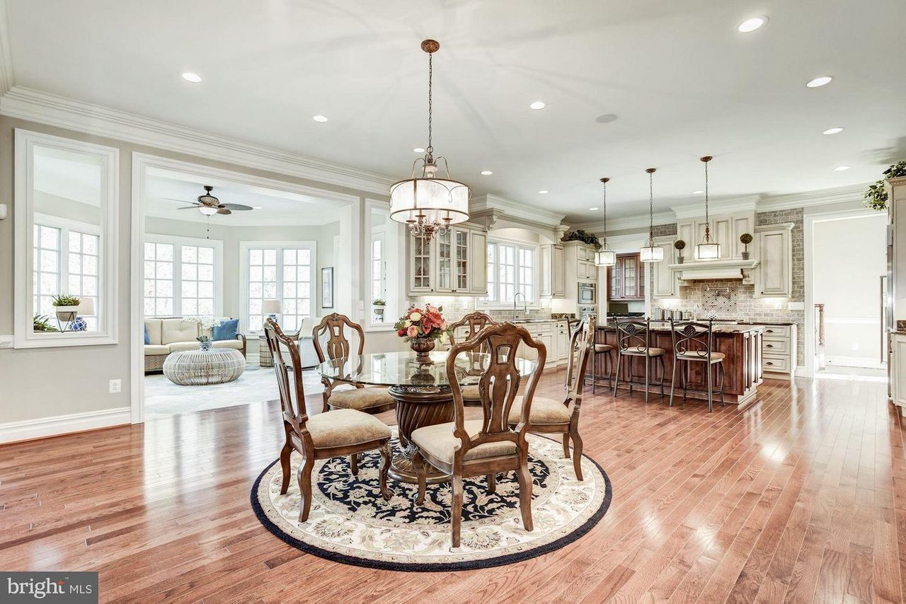 Spacious kitchen and dining area with wooden floors, round glass dining table, ornate chairs, kitchen island with stools, and pendant lighting. Large windows and cozy seating area in the background.