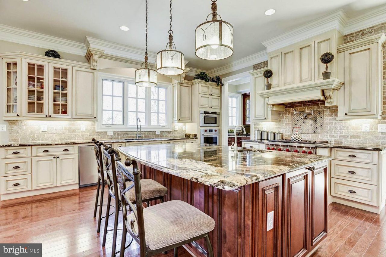 Spacious kitchen with cream cabinets, granite island, pendant lights, and stainless steel appliances. Hardwood floors and tiled backsplash complete the look.