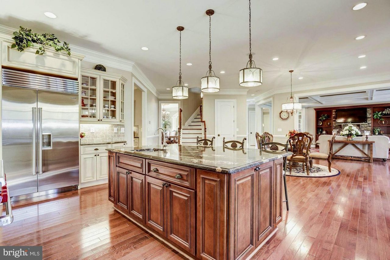 Spacious kitchen with wooden floors, an island with dark marble countertop, stainless steel appliances, hanging lights, and a view of a living area with a dining set.