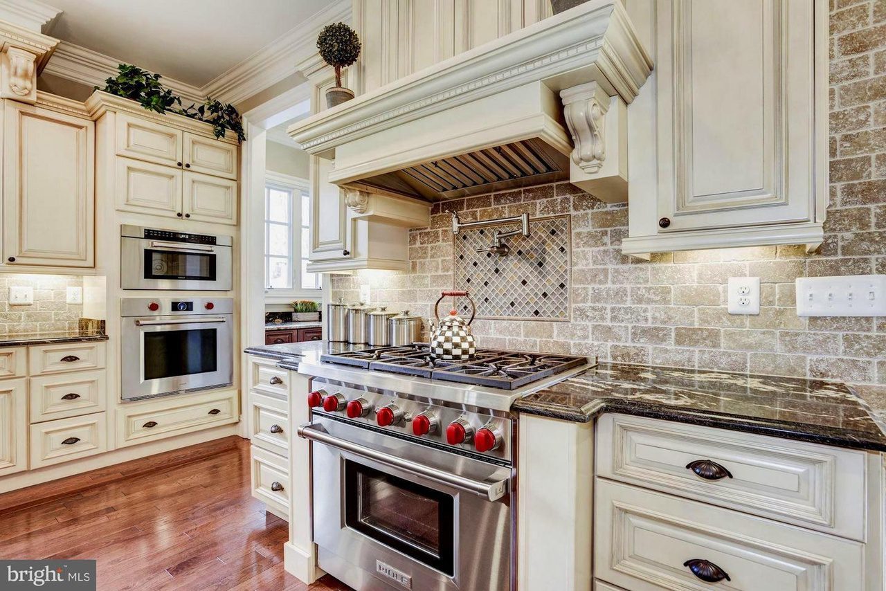 A kitchen with cream cabinets, brick-patterned backsplash, stainless steel stove with red knobs, and a checkered teapot on the burner. Nearby are a built-in oven and a microwave.