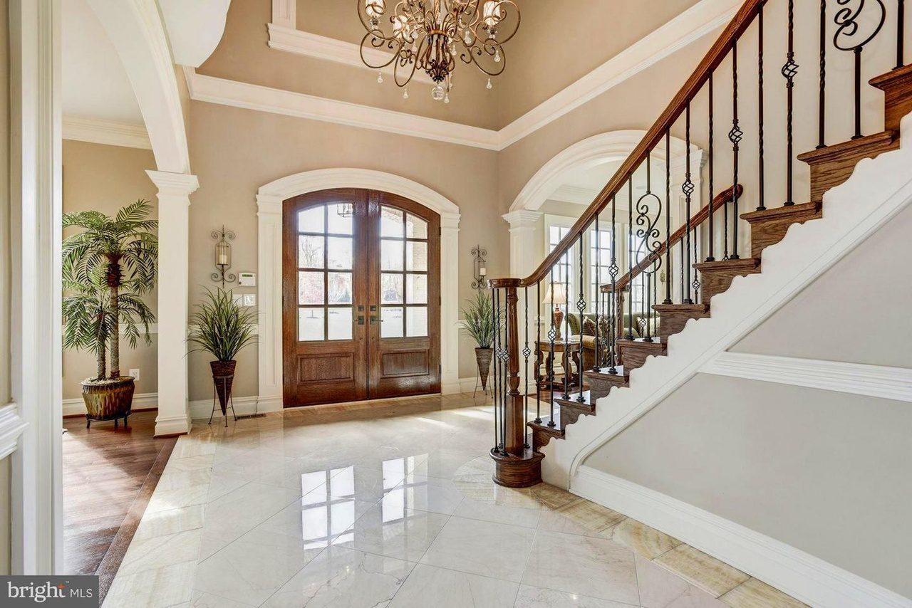 Elegant foyer with a polished marble floor, a grand wooden staircase, and a chandelier. Double doors flanked by potted plants enhance the space's sophistication.