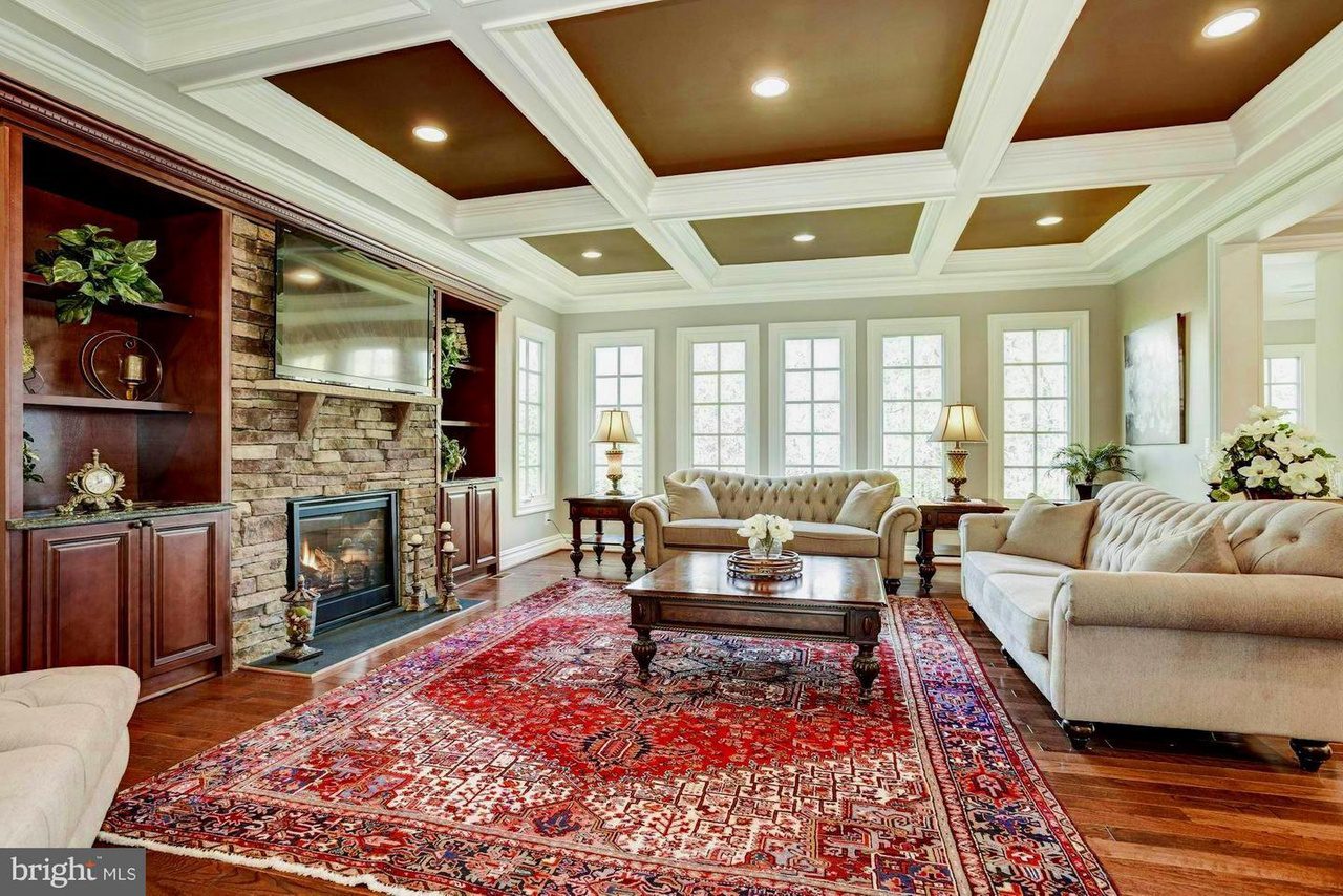 A living room with a coffered ceiling, large windows, two beige sofas, a fireplace with a TV above, wooden built-in shelves, and a red patterned rug on wood flooring.