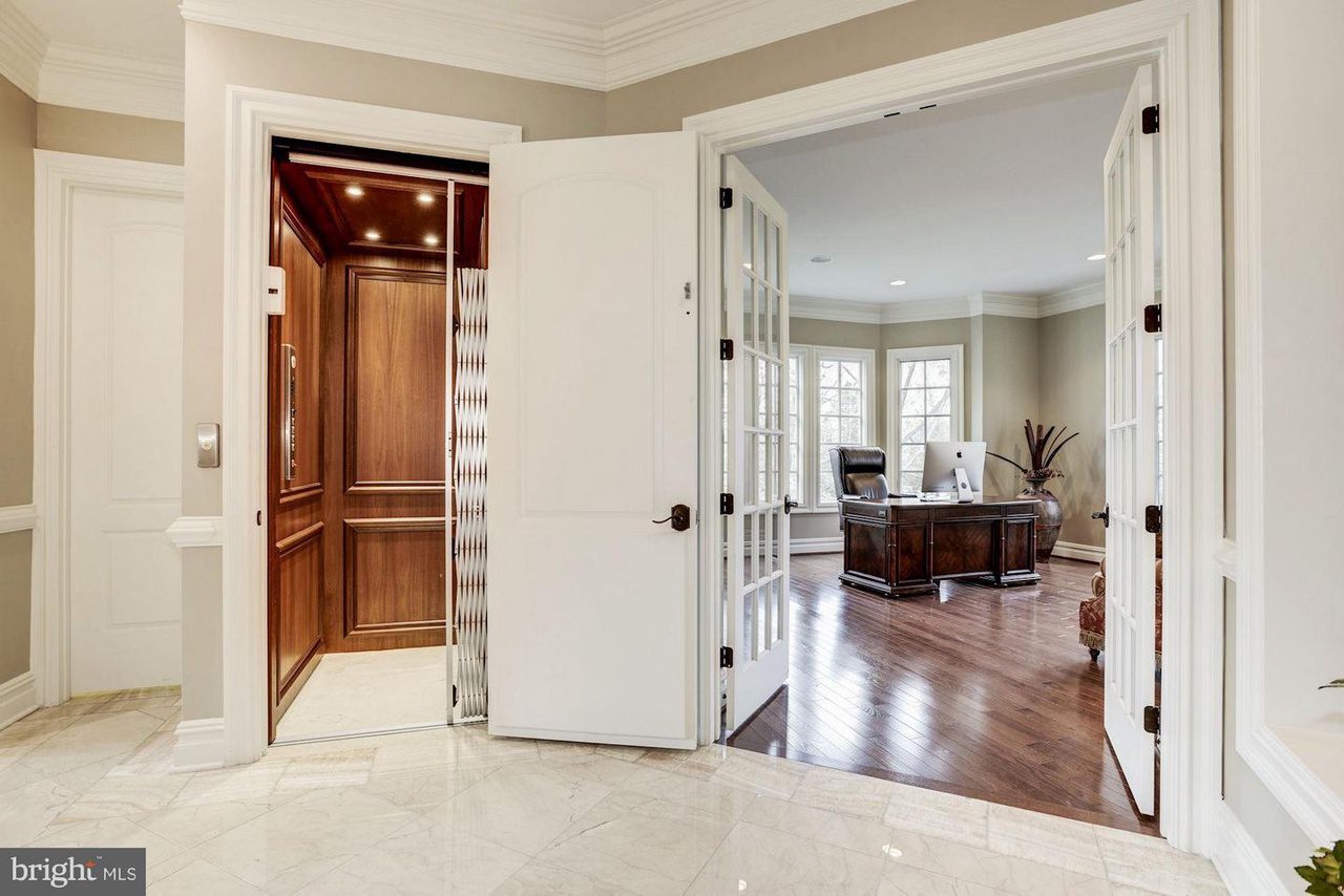 Elegant home interior with office and open elevator. Office features wooden desk, armchair, and large windows. Elevator shows wood paneling and metal gate. Marble tiles in hallway.