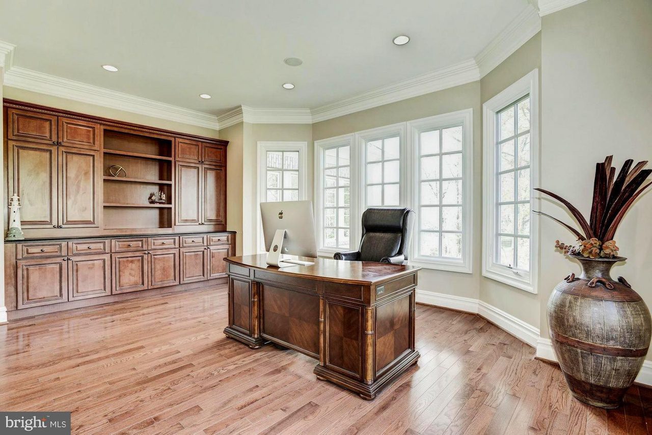 Spacious office with a large wooden desk, leather chair, built-in wooden shelving, and floor-to-ceiling windows. Decor includes a decorative vase and various items on the shelves.