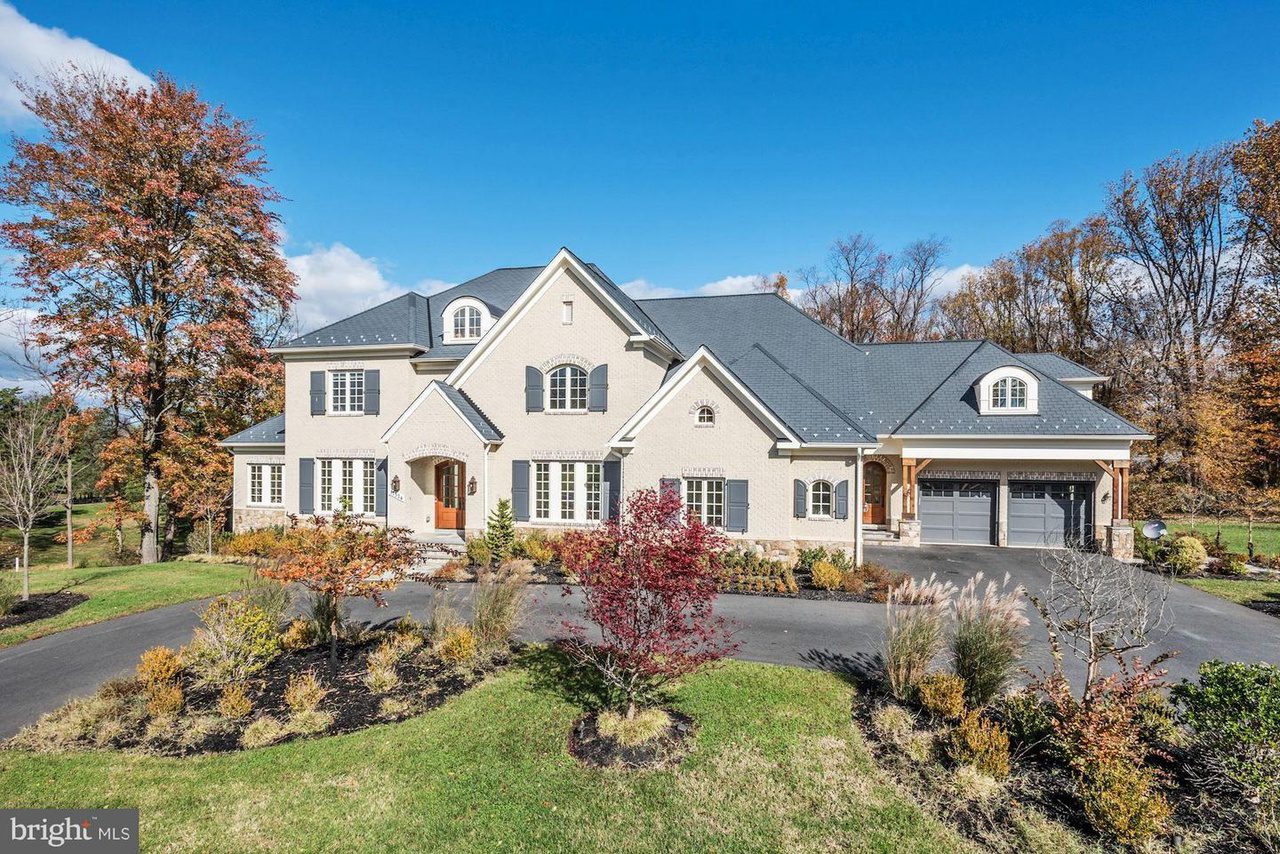 Large two-story house with beige exterior and dark roof, surrounded by landscaped lawn and autumn trees. Attached three-car garage is visible on the right. Clear blue sky overhead.