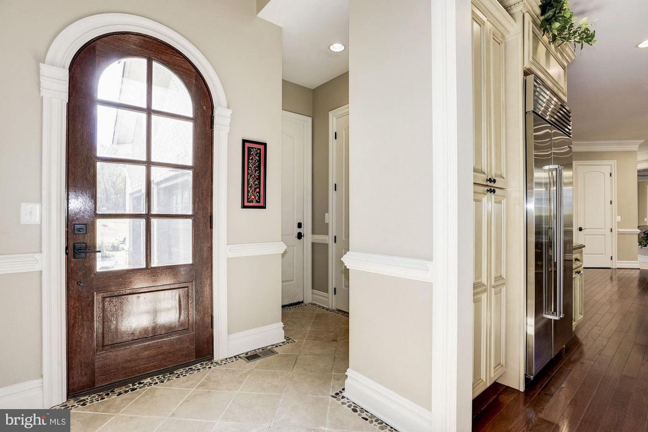 A hallway with beige tile flooring, a large wooden arched door with glass panels, and a stainless steel refrigerator next to light cream cabinets.