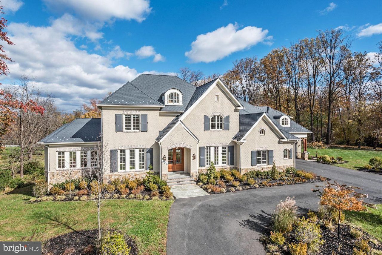 Large two-story house with gray roof and beige exterior, surrounded by landscaped garden and trees under a partly cloudy sky.