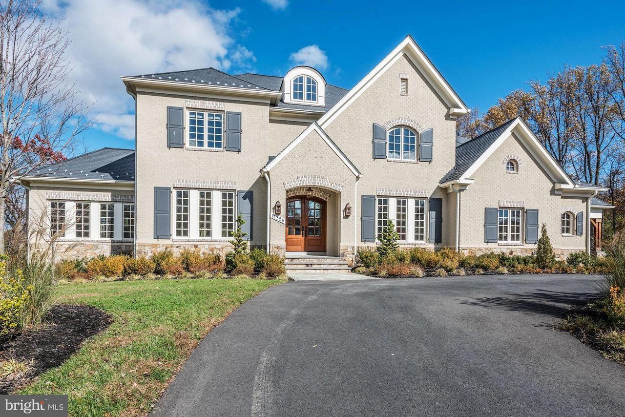 Large two-story house with beige exterior, blue shutters, and a circular driveway, surrounded by trees and shrubs.