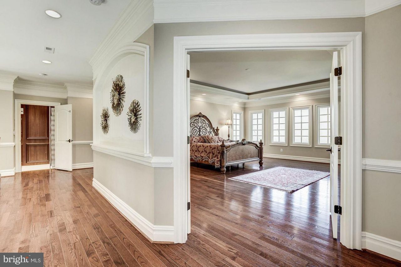 Spacious hallway with wooden flooring leading to an ornate bed in a bedroom with large windows and a decorative rug.