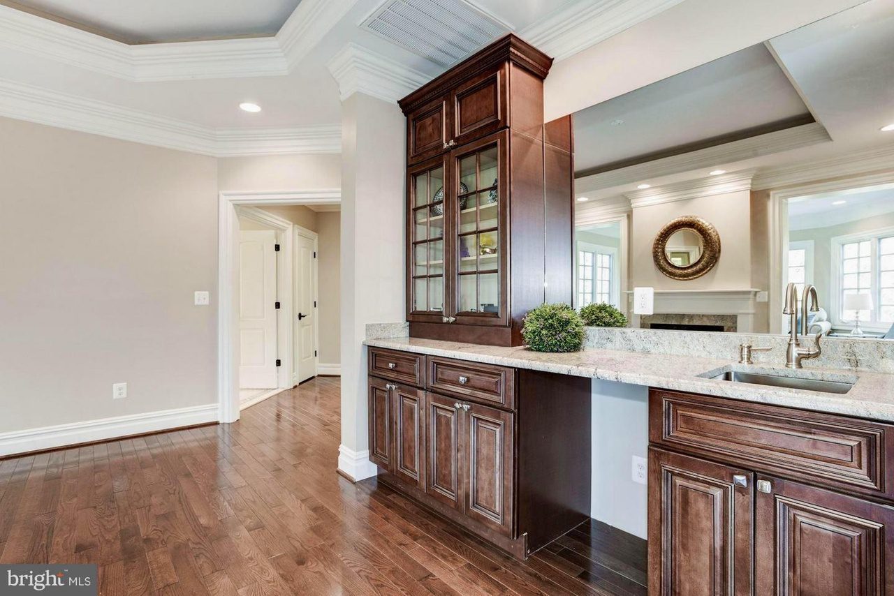Kitchen area with dark wood cabinets, glass-paneled doors, granite countertops, and a sink. There's an open doorway on the left and a mirror with a circular frame in the background.