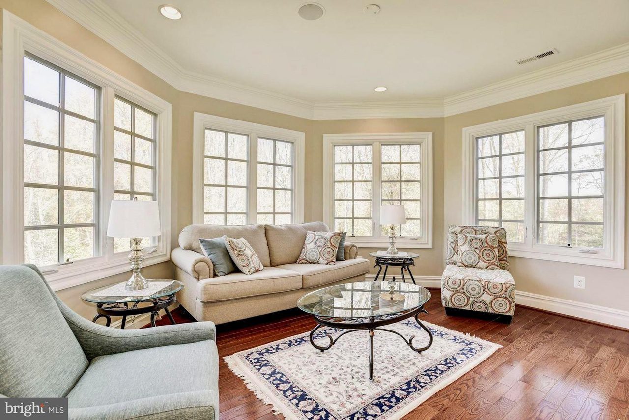 A sunlit room with beige walls features a sofa, two armchairs, and a glass coffee table on a patterned rug. Large windows surround the area, providing natural light.