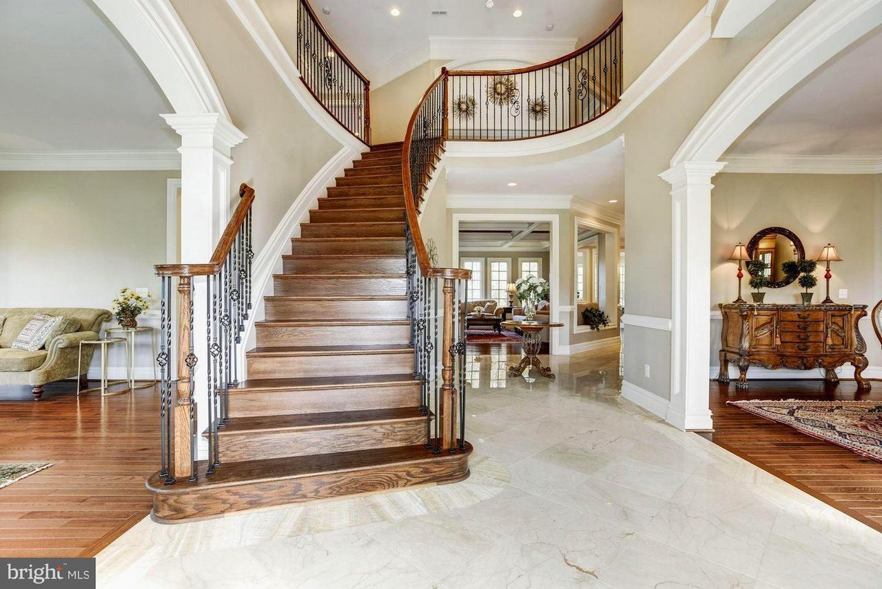 Elegant, curved wooden staircase with wrought iron railings in a spacious foyer, featuring polished marble flooring, cozy seating area, and decorative furniture.