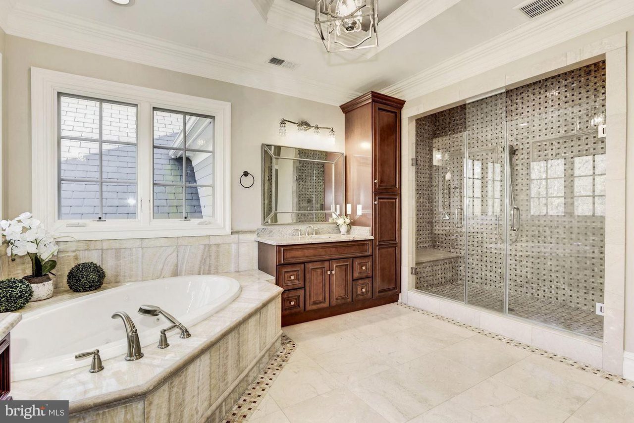 Luxury bathroom with a corner bathtub, glass-enclosed shower, dark wood vanity, and large mirror. A window is on the left wall, and there are decorative plants beside the tub.