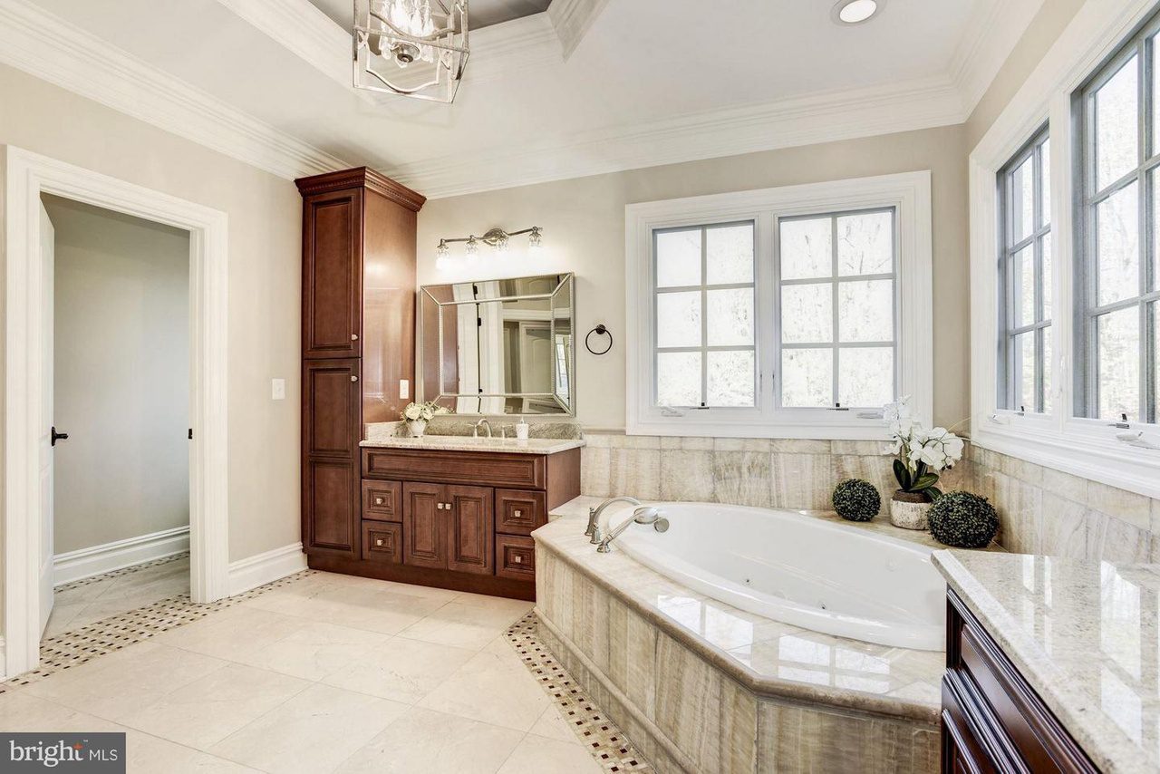 Luxurious bathroom with large corner bathtub, wood vanity with mirror, dual window, tile flooring, and beige walls.