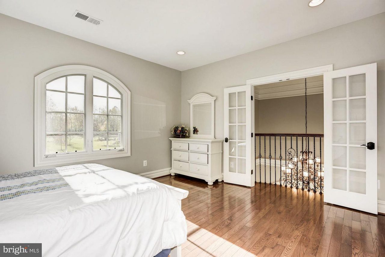 A sunlit bedroom with a bed, arched window, white dresser, and French doors opening to a balcony with an iron railing. Hardwood floor and neutral walls complete the space.