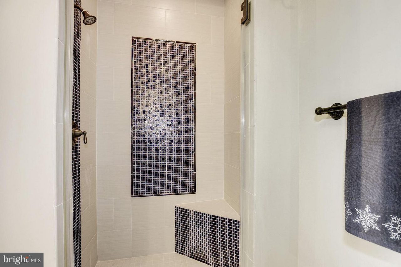 A shower with white tile walls, a mosaic accent of blue and black tiles, a built-in corner seat, and a towel with snowflake design on a nearby rod.